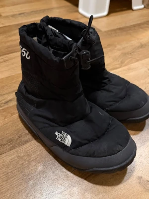 Svarta vinterboots från The North Face - Säljer ett par svarta vinterboots från The North Face i storlek 38. Boots med vadderad ovandel, dragsko upptill och slitstark sula. Perfekta för kalla dagar och riktigt sköna att ha på sig. Klassisk logga på sidan och enkel, sportig design.