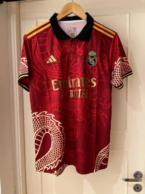 Real Madrid red dragon special edition jersey - Säljer en limited edition Real Madrid x Adidas matchtröja i rött med guldiga detaljer och ett stort drak soprint över hela tröjan. Klassisk svart krage, gula axelränder och broderad klubbmärke. Slim fit och tillverkad i lätt polyester, perfekt för dig som vill sticka ut.