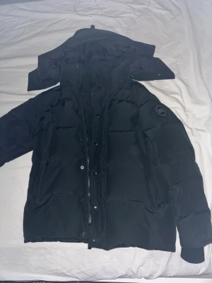 Svart dunjacka från Canada Goose - Säljer en äkta svart dunjacka från Canada Goose med avtagbar huva och klassisk patch på ärmen. Jackan har dragkedja och knappar framtill, stora fickor och är perfekt för kalla vinterdagar. Fodret är glansigt och jackan har en stilren, enkel design. Väskan medföljer.