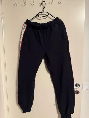 Svarta mjukisbyxor med röd/vit detalj - Svarta mjukisbyxor med elastisk midja och mudd vid benslut. Sidorna har en röd och vit rand med text och stjärnor, vilket ger en sportig vibe. Perfekta för chill eller träning. Snygg och bekväm passform. 