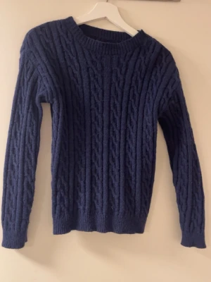 Mörkblå kabelstickad tröja - Säljer en mörkblå kabelstickad tröja med rund hals och långa ärmar. Tröjan har ett klassiskt flätmönster och ribbade muddar vid ärmslut och nederkant. Perfekt för kyliga dagar och enkel att styla till olika outfits.