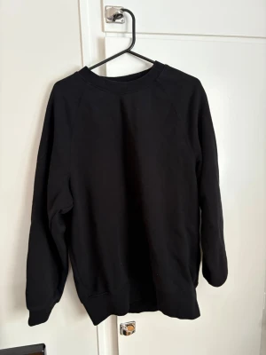 Svart sweatshirt i bomull - En stilren svart sweatshirt med rund hals och långa ärmar. Tröjan har en klassisk passform och är tillverkad i mjuk bomull, perfekt för att chilla eller hänga med kompisar. Ribbstickade muddar vid ärmslut och nederkant ger en snygg look.