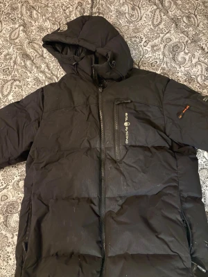 Svart pufferjacka från Sail Racing - Säljer en svart pufferjacka från Sail Racing med huva och dragkedja framtill. Jackan har en bröstficka med dragkedja och logga, samt en ficka på ärmen med orange detalj. Perfekt för kalla dagar och riktigt skön att bära.