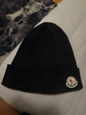Svart Moncler mössa  - Säljer en svart ribbstickad mössa från Moncler med klassisk logga framtill. Mössan är tillverkad i mjuk ull och har en vikt kant för extra stilpoäng. Perfekt accessoar för kalla dagar och snygg till alla outfits.