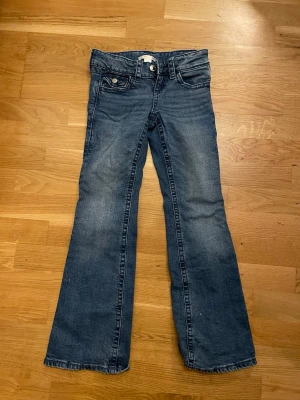 Blå bootcut jeans från Gina Tricot Young - Snygga blå jeans från Gina Tricot i bootcut-modell med coola paljettrosetter på bakfickorna. Klassisk femficksdesign, knapp och dragkedja fram. Jeansen är gjorda i jeansmaterial och har en normal passform. Perfekt för dig som gillar detaljer och vill sticka ut lite extra.