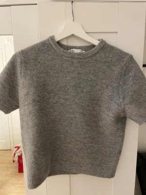 Grå stickad tröja från Zara - Mysig grå stickad (lite kortare) tröja från Zara med korta ärmar och rund hals. Tröjan har en enkel och clean design med ribbade kanter vid hals, ärmslut och nederkant. Perfekt för lager-på-lager eller att ha i sig självt. Använd fåtal gånger i fint skick!💕
