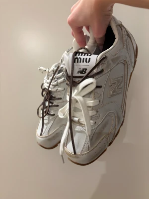 Miu Miu x New Balance - Unika vita sneakers från Miu Miu x New Balance med bruna och vita snören, klassisk NB-logga på sidan och cool kontrast mellan skinn och mockadetaljer. Snygg gummisula och detaljerad design som sticker ut. Perfekta för dig som vill ha något extra. Ej autentiska