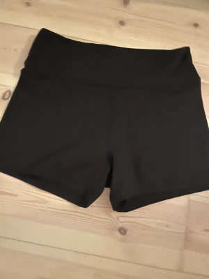 Svarta träningsshorts från Move - Svarta, högmidjade träningsshorts från Move med stilren design och kort passform. Perfekta för gymmet eller löprundan, tillverkade i mjukt och stretchigt syntetmaterial som sitter skönt mot kroppen. Diskret logga framtill i midjan.