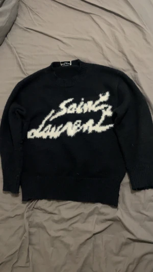 Svart stickad Saint Laurent tröja - Svart stickad tröja från Saint Laurent med vit, fluffig logotyp framtill. Rund hals och långa ärmar. Perfekt för dig som gillar statement-plagg och vill ha något som sticker ut i garderoben.