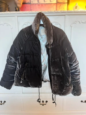 Svart pufferjacka från Odd Molly - Säljer en svart pufferjacka från Odd Molly med mjuk, glansig yta i sammet. Jackan har hög krage, två stora fickor med dragkedja och snörning nedtill. Insidan har ett snyggt mönstrat foder och ett broderat Odd Molly-märke på ärmen. Perfekt för dig som vill ha en cool och mysig jacka.