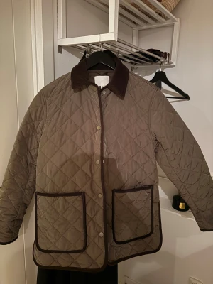 Quiltad brun jacka från H&M - Säljer en quiltad brun jacka från H&M med mörkbrun krage och kantband. Jackan har stora framfickor och tryckknappar framtill. Perfekt för dig som gillar klassisk stil med en modern twist.