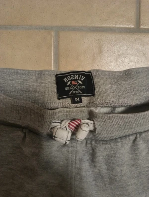 Grå mjukisbyxor Vinson Polo Club - Grå mjukisbyxor från Vinson Polo Club med snörning i midjan och muddar vid bensluten. Byxorna har tryck med siffra och logga framtill samt fickor på sidorna och en bakficka. Loggan är lite sudding. Perfekta för chill eller träning.