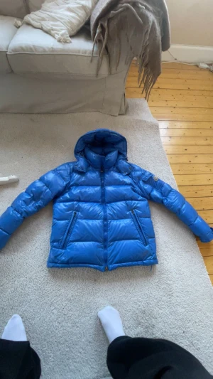 Blå Moncler pufferjacka med huva - Säljer en blå pufferjacka från Moncler med huva och dragkedja framtill. Jackan har två sidofickor med dragkedja och klassisk Moncler-logga på ärmen. Materialet är glansigt och fyllt med dun för extra värme. Perfekt för dig som vill ha en snygg och varm jacka i vinter.