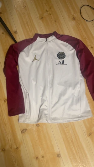 PSG x Jordan vit och vinröd track jacket - Snygg track jacket från PSG x Jordan med vit kropp och vinröda ärmar. Jackan har dragkedja framtill, guldigt Jumpman-logo och PSG-märke på bröstet. Perfekt för dig som älskar fotboll och streetwear.