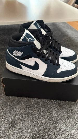 Nike Air Jordan 1 Mid Navy/Vit - Säljer ett par Nike Air Jordan 1 Mid i marinblått och vitt med svarta skosnören. Klassisk siluett med perforerad tå, vit logga på sidan och mjuk vadderad plös. Ovandel i skinn och syntet, perfekt för dig som gillar streetwear och sneakers med ikonisk design. Kvitto finns! 