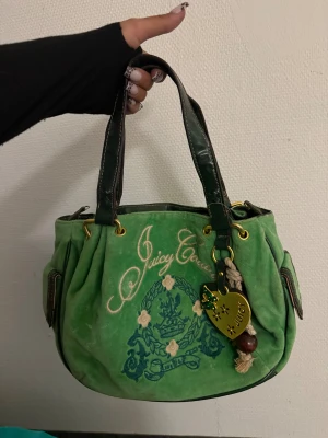 Grön handväska Juicy Couture  - Snygg grön handväska från Juicy Couture i mjuk manchester med broderad logga och blommor framtill. Väskan har mörkgröna detaljer, guldfärgade metalldelar och en stor hjärtformad Juicy-tag med fjäril. Insidan är fodrad med rosa tyg och har innerfack med dragkedja.