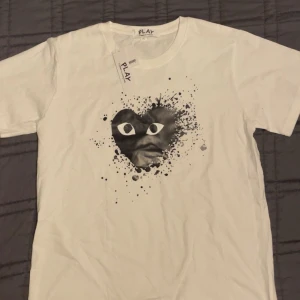 Vit t-shirt Comme des Garçons Play (prisförslag)  - Snygg vit t-shirt från Comme des Garçons Play i storlek XL. Sitter som en LFramsidan har ett svart hjärta med ögon och coolt splattertryck. Klassisk rund hals och korta ärmar. Materialet är mjuk bomull, perfekt för en chill och trendig look.
