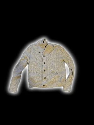 Grå stickad kofta med knappar - Mysig grå stickad kofta med grovt kabelmönster och stora svarta knappar framtill. Koftan har två fickor och en hög krage som ger extra värme. Perfekt för kyliga dagar och passar dig som gillar klassisk stil med en modern twist.