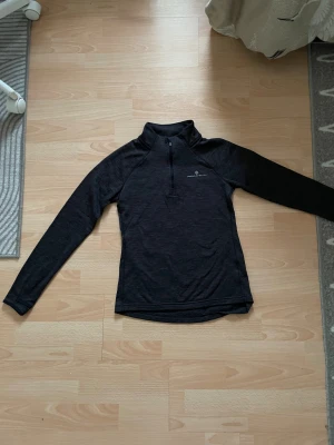 Svart långärmad träningströja från Ronhill - Sportig svart långärmad träningströja från Ronhill med half zip framtill. Tröjan har en smal passform och är tillverkad i mjukt syntetmaterial som andas. Perfekt för löpning eller gym. Diskret logga på bröstet och hög krage för extra komfort. Står dock ingen storlek på den men skulle säga ungefär storlek S