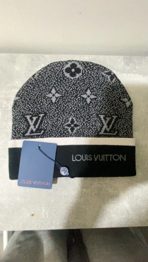 Svart och vit Louis Vuitton mössa - Snygg svart och vit mössa från Louis Vuitton med klassiskt LV-monogram och blommönster. Mössan har en bred vit kant och logotypen broderad framtill. Materialet är stickat och ger en mjuk och skön känsla. Perfekt för dig som vill ha en lyxig och trendig look.