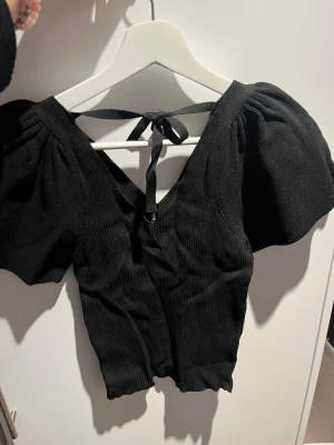 Svart ribbad topp med puffärm och knyt - Svart ribbad topp med korta puffärmar och snygg v-ringning. Toppen har ett knytband i ryggen som ger en extra detalj och sitter croppad. Materialet är mjukt och stretchigt, perfekt för att skapa en cool look. Passar dig som gillar statement-ärmar och trendiga detaljer.
