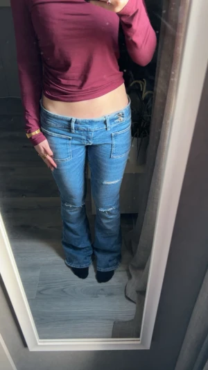 Lågmidjade bootcut jeans med hål - Säljer ett par blå jeans med låg midja och bootcut-ben. Jeansen har hål på låren och knäna, samt dubbla knappar i midjan. Klassisk femficksmodell i mjuk denim som sitter snyggt över höfterna.