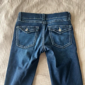 Mörkblå bootcut jeans med kontrastsömmar - Snygga mörkblå jeans med bootcut passform och tydliga vita kontrastsömmar. Klassisk femficksmodell med knappar på bakfickorna och detaljerade sömmar. Perfekta för dig som gillar en tidlös och cool look.