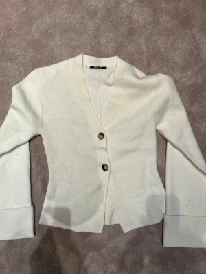 Vit kofta från Gina Tricot med knappar - Snygg vit kofta från Gina Tricot med två stora bruna knappar framtill. Koftan har långärmad design och breda muddar vid ärmsluten. Materialet känns mjukt och är troligtvis bomull eller polyester. Perfekt för lager-på-lager under kyliga dagar.