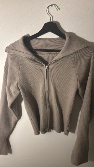 Beige ribbad kofta med dragkedja - Snygg beige ribbad kofta med dragkedja framtill och bred krage. Långärmad modell med tumhål i ärmsluten för extra chill vibe. Perfekt att slänga på sig när det är lite kyligt ute. Materialet känns mjukt och stickat, vilket gör den både bekväm och stilren. Passar både XS och S