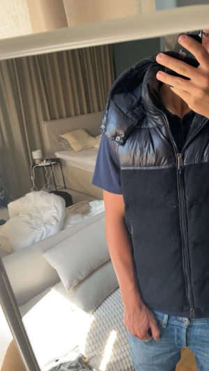 Svart dunväst från Polo Ralph Lauren - Svart dunväst från Polo Ralph Lauren med glansig dunpanel upptill och mjukare tyg nedtill. Västen har huva, två dragkedjefickor och hel dragkedja framtill. Perfekt för lager-på-lager och snygg till jeans eller hoodie.