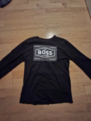 Svart långärmad tröja från Boss - Svart långärmad tröja från Boss med stort tryck på bröstet i vitt och grått. Tröjan har en klassisk rund hals och är gjord i mjuk bomull som känns skön mot huden. Perfekt för dig som gillar stilrena och enkla plagg med tydlig logga. Barn storlek S