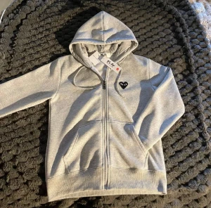 Hoodie Comme des Garçons Play - Grå hoodie med dragkedja från Comme des Garçons Play. Tröjan har huva med snörning, två fickor fram och ett svart hjärta med ögon på bröstet samt ett stort hjärta med ögon på ryggen. Tillverkad i mjuk bomull, perfekt för chill dagar. PRIS KAN DISKUTERAS