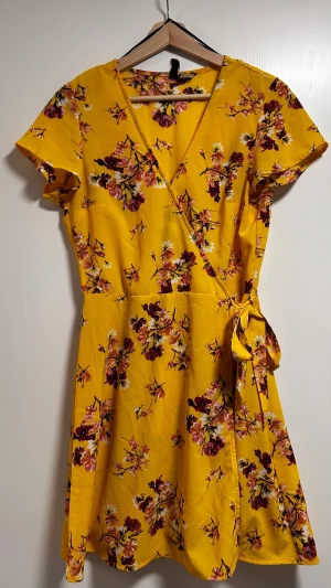 Gul blommig omlottklänning - Superfin gul omlottklänning från H&M med blommigt mönster i rosa, vinrött och vitt. Klänningen har korta ärmar, v-ringning och knyt i midjan. Perfekt för sommarens alla tillfällen och ger en färgglad vibe.
