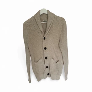 Beige stickad kofta med sjalkrage - Säljer en beige stickad kofta från selected med sjalkrage och stora svarta knappar framtill. Koftan har två praktiska fickor och en grovstickad struktur som ger en chill och mysig vibe. Perfekt att slänga på sig när det är lite kyligt ute.