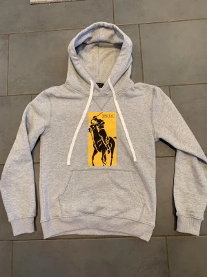 Grå hoodie Polo Ralph Lauren M - Snygg grå hoodie från Polo Ralph Lauren i storlek M. Tröjan har en stor gul patch med svart polospelare på bröstet, vita snören och klassisk känguruficka. Mjuk bomullsmix och huva gör den extra chill. Perfekt för dig som gillar sportig och stilren streetwear. Påsen och taggarna kommer dock inte med på köpet