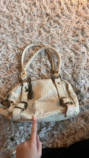 Beige handväska från Friis & Company - Snygg beige handväska från Friis & Company med flätad look och coola metalldetaljer i antik mässing. Väskan har dubbla handtag, dekorativa ringar och charmiga hängen. Perfekt storlek för dina essentials och har dragkedja på insidan.