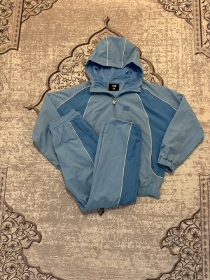 Blå Tracksuit från FSBN med huva - Säljer en ljusblå träningset från FSBN med huva och dragkedja. Jackan har vita detaljer längs ärmarna och sidorna, samt elastiska muddar vid ärmslut och nederkant. Perfekt för en sportig look!🩵 Byxorna är Storlek XS och Jackan är S men byxorna blir som ett S eftersom den är baggy. Priset är inte i hugget, kan diskutera 