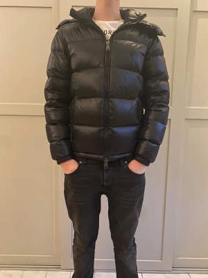 Svart dunjacka från Ralph Lauren - Säljer en svart, glansig dunjacka från Ralph Lauren med huva och dragkedja framtill. Jackan har klassisk pufferstil, broderad logga på bröstet och är riktigt varm och skön för kalla dagar. Perfekt för dig som vill ha en stilren och sportig look. Jackan passar alltifrån 174-185 cm. Jackan är använd ca 10 gånger. Den är nästintill ny och har inga defekter. Nypris är ca 6500 kr! 