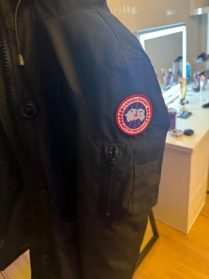 Svart vinterjacka  från Canada Goose - Svart parkajacka från Canada Goose med klassisk patch på ärmen, dragkedja och knappar framtill samt justerbar huva. Luvan har lite smink på sig och drar ner priset på grund av det.