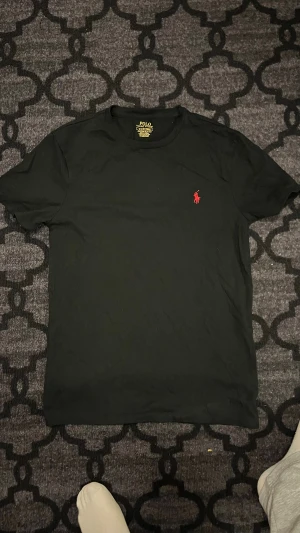 Svart t-shirt Polo Ralph Lauren - Svart t-shirt från Polo Ralph Lauren med röd broderad logga på bröstet. Modellen är custom slim fit och har rund hals samt korta ärmar. Materialet är mjuk bomull som känns skön mot huden. Perfekt för dig som gillar stilrena och klassiska plagg.