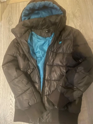 Svart pufferjacka med blått foder - Svart pufferjacka med blått innerfoder och huva. Jackan har quiltad design, ribbade muddar och dragkedja framtill. Märket 99 broderat på bröstet i blått. Perfekt för kyliga dagar när du vill hålla stilen och värmen.