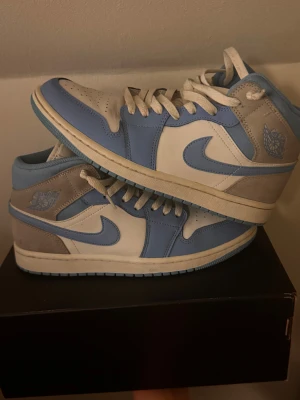 Nike Air Jordan 1 Mid UNC Blå/Vit - Nike Air Jordan 1 Mid UNC i fräscha blå och vita färger med grå mockadetaljer. Klassisk siluett med snörning, rund tå och platt sula. Swoosh-logga på sidan och Air Jordan Wings på ankeln. Perfekt för dig som vill ha en ikonisk sneaker med snygg färgkombo.