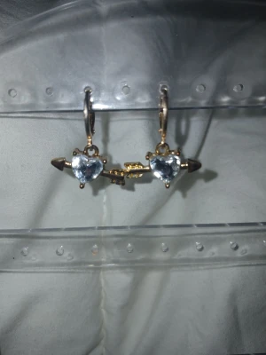 Hjärtformade örhängen med pil - Supersöta örhängen i guldton med glittrande hjärtformade stenar i silverfärg. Varje örhänge har en pil som går genom hjärtat, vilket ger en unik och lekfull look. Perfekta för att addera lite extra bling till din outfit.