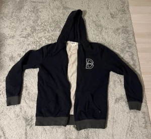 Mörkblå hoodie från Dobber med dragkedja - Snygg mörkblå hoodie från Dobber i storlek medium. Tröjan har huva med snörning, dragkedja framtill och två fickor. Ribbstickade muddar vid ärmslut och nederkant. Stor D-logga på bröstet ger en cool streetkänsla.