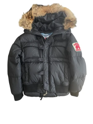 Svart dunjacka från Dsquared2 med päls - Svart dunjacka från Dsquared2 med stor huva med äkta päls. Jackan har flera fickor framtill, ribbade muddar och patch på ärmen. p2p 56  längd framsida 63 längd baksida 70
