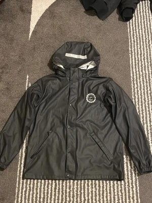 Svart regnjacka från Tuxer med huva - Svart regnjacka från Tuxer med huva och reflexdetaljer. Jackan har två snedställda fickor framtill, tryckknappar och dragkedja. Materialet är syntet och jackan har en vit insida. Perfekt för regniga dagar när du vill hålla dig torr.