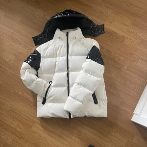 Moose Knuckles puffjacka - Säljer en fet pufferjacka från Moose Knuckles i vit med svarta detaljer och glansiga partier på ärmar, rygg och huva. Jackan har dragkedja, hög krage och stor logga på ärmen. Perfekt för kalla dagar när du vill sticka ut.