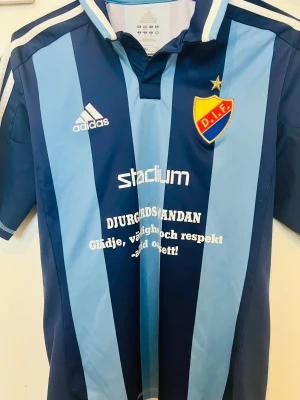 Djurgården fotbollströja Adidas - Djurgården IF matchtröja från Adidas i polyester med klassiska breda ljusblå och mörkblå ränder, broderat DIF-emblem med stjärna, tryckt nummer 8 på ryggen och vita Adidas-loggan på bröstet. Snygg krage och tryck från Stadium på framsidan.