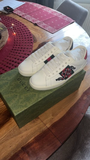 Gucci Ace sneakers - Säljer ett par vita Gucci Ace sneakers i skinn med klassiska gröna och röda ränder på sidan och broderad orm. Baksidan har metallicröd och metallicgrön detalj med Gucci-logga. Skorna har snörning och platt sula, samt rund tå. Riktigt snygga och ikoniska sneakers för dig som vill sticka ut.