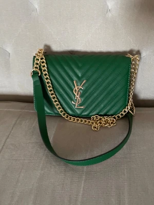 Grön quiltad axelväska från YSL - Snygg grön axelväska från YSL med guldfärgad kedja och logga framtill. Väskan har quiltad design, magnetlås och insida i rött med flera fack och dragkedja. Perfekt storlek för det viktigaste och ger en lyxig vibe till din outfit.